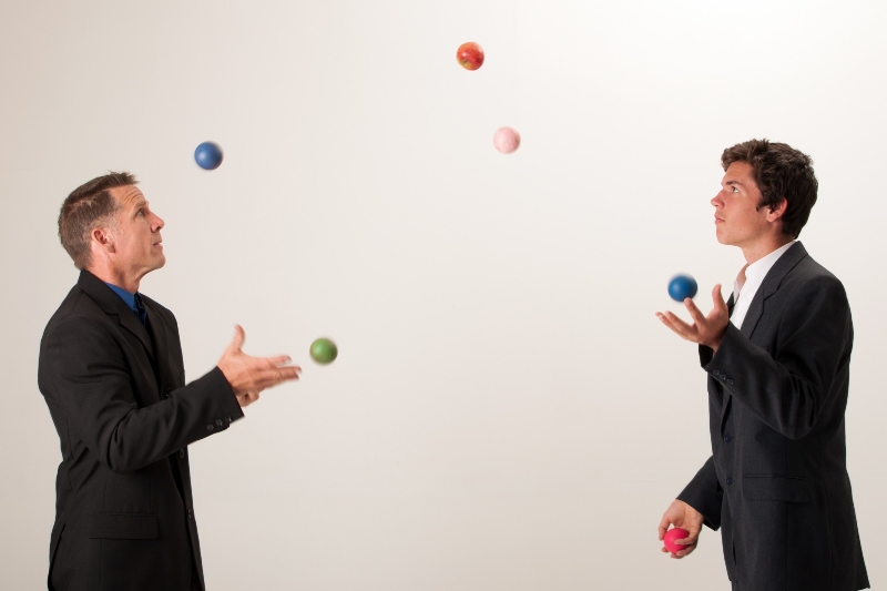 two businessmen juggle numerous brightly colored balls and one red apple